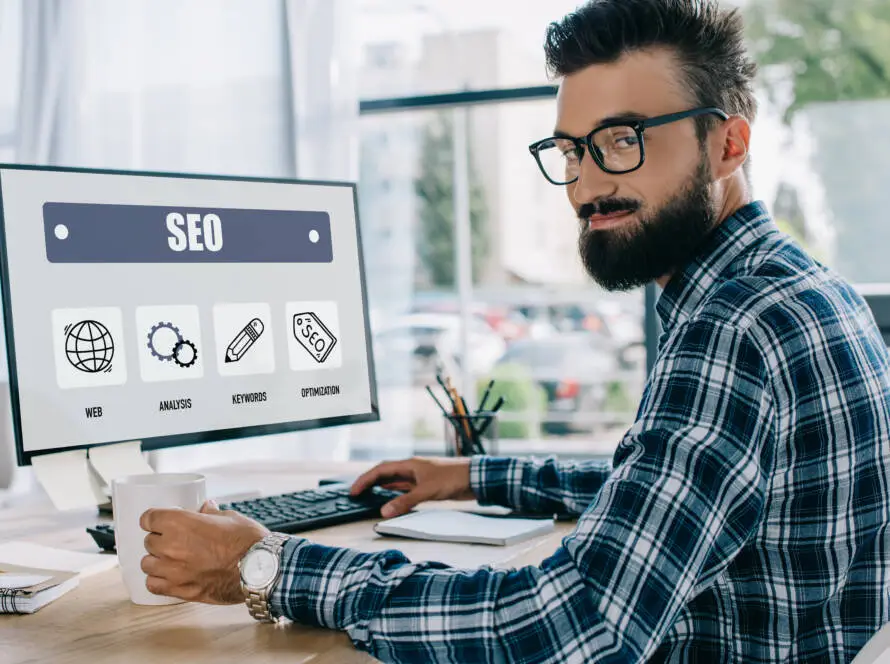 A bearded man in glasses and a checked shirt sits at a desk, holding a mug and using a computer. The monitor displays an SEO dashboard, suggesting he could be a healthcare SEO consultant working in a bright office with large windows.