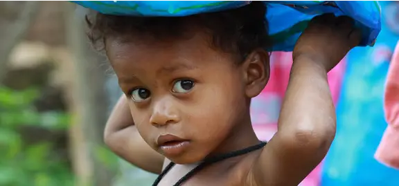 A young child with curly hair looks towards the camera, holding a blue sheet above their head for shade. The photo’s natural setting could inspire a healthcare digital marketing agency seeking authentic, relatable imagery for campaigns.