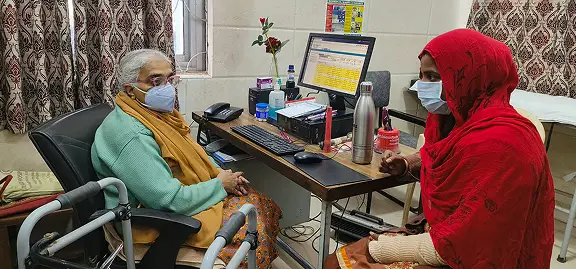 An elderly woman with a zimmer frame and a younger woman in a red headscarf, both wearing masks, sit opposite each other in a doctor's surgery—a scene reflecting the trust vital for effective digital marketing healthcare strategies.
