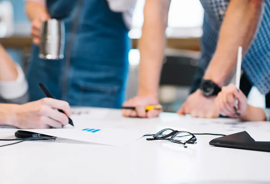 Close-up of a group collaborating at a table—hands writing and pointing on paper with pens and markers. Spectacles, a folded black case, and a metallic coffee mug rest on the white table—ideal for a digital marketing healthcare team brainstorming ideas.