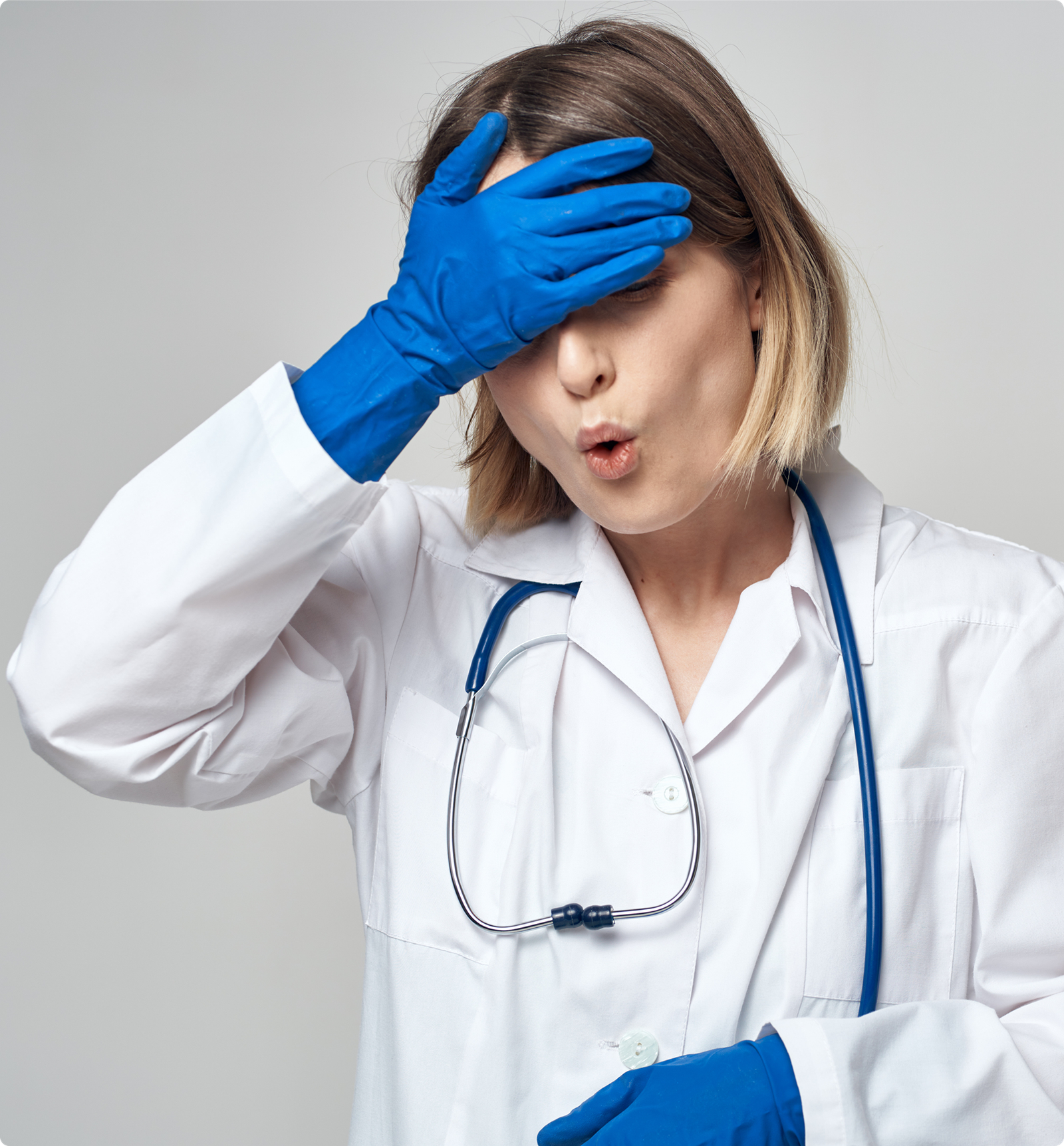 A woman in a white lab coat and blue gloves, with a stethoscope round her neck, covers her forehead with one hand and makes a surprised, “oops” expression. She has straight, chin-length blonde hair and stands against a plain light background.