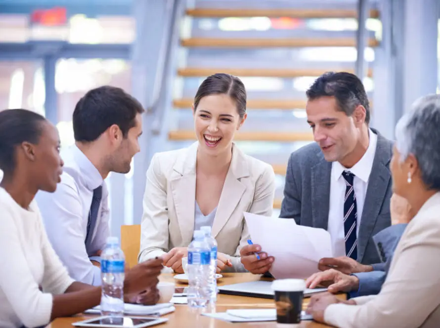 Five professionals sit around a table in a Healthcare Marketing Agency office, engaged in lively discussion. Three women and two men smile and interact, holding papers and pens. Water bottles, a coffee cup, and tablets are on the table.