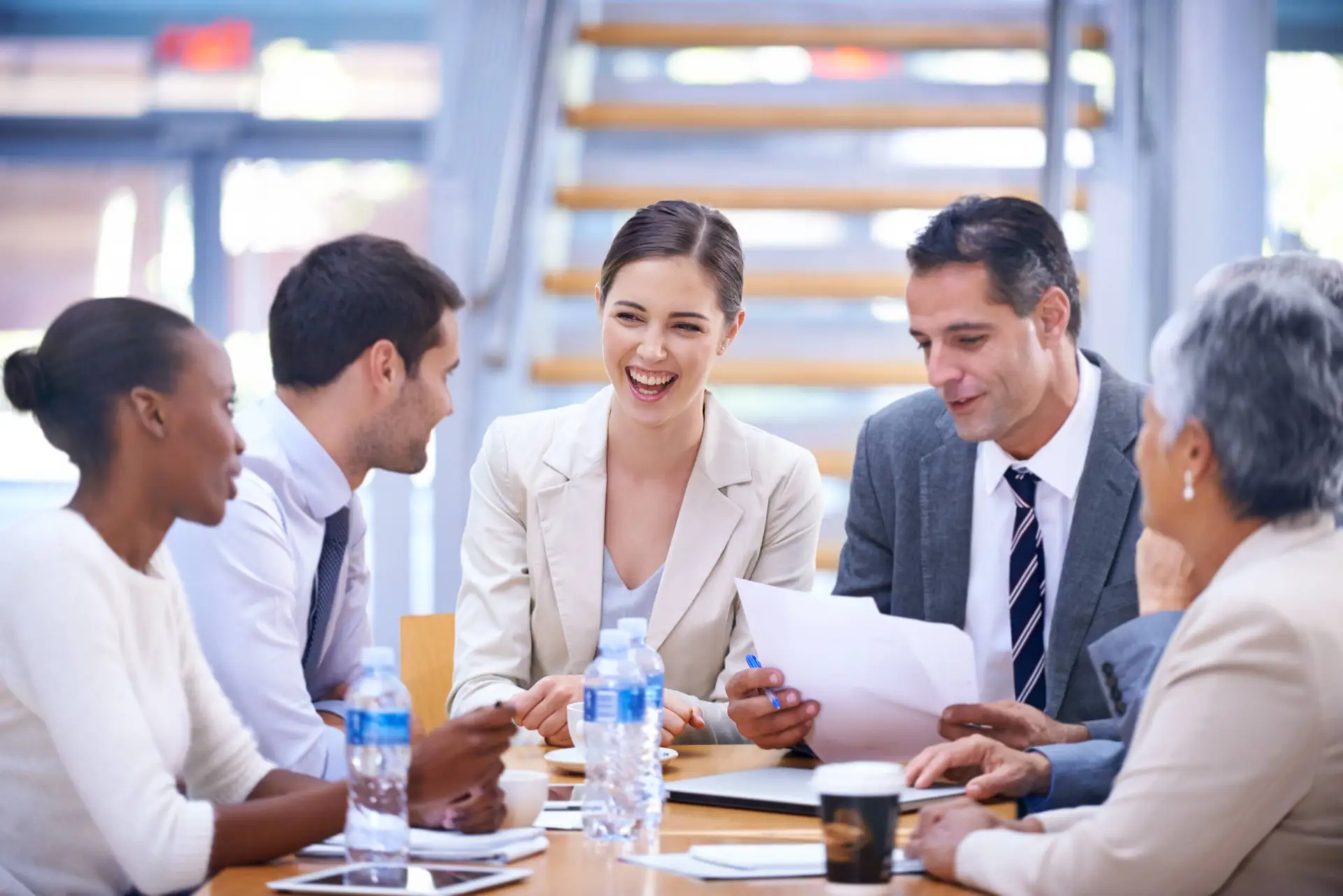 Five professionals sit around a table in a Healthcare Marketing Agency office, engaged in lively discussion. Three women and two men smile and interact, holding papers and pens. Water bottles, a coffee cup, and tablets are on the table.