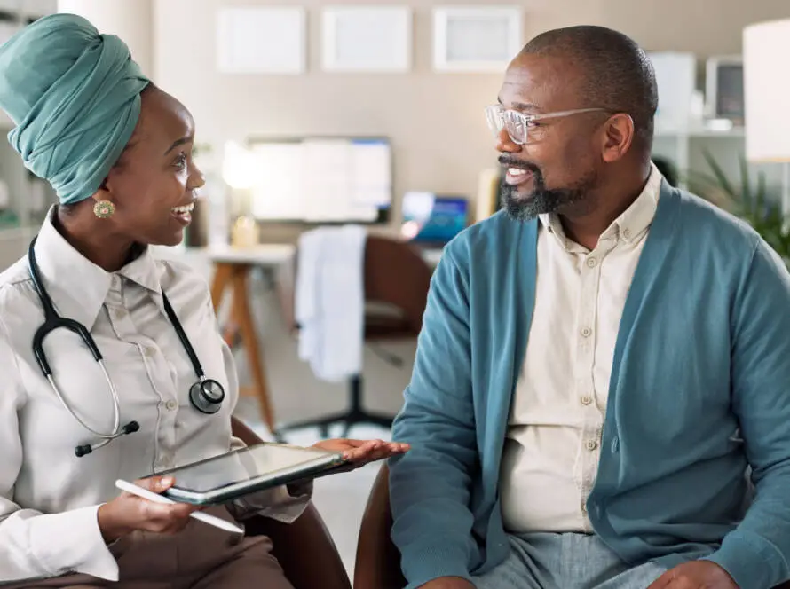 A female doctor in a white coat and headscarf holds a digital tablet, chatting with a male patient in glasses and a blue cardigan. Both are smiling in a bright, modern medical surgery—an ideal scene for any healthcare marketing agency campaign.