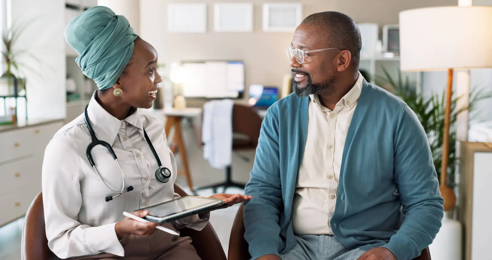 A female doctor in a white coat and headscarf holds a digital tablet, chatting with a male patient in glasses and a blue cardigan. Both are smiling in a bright, modern medical surgery—an ideal scene for any healthcare marketing agency campaign.