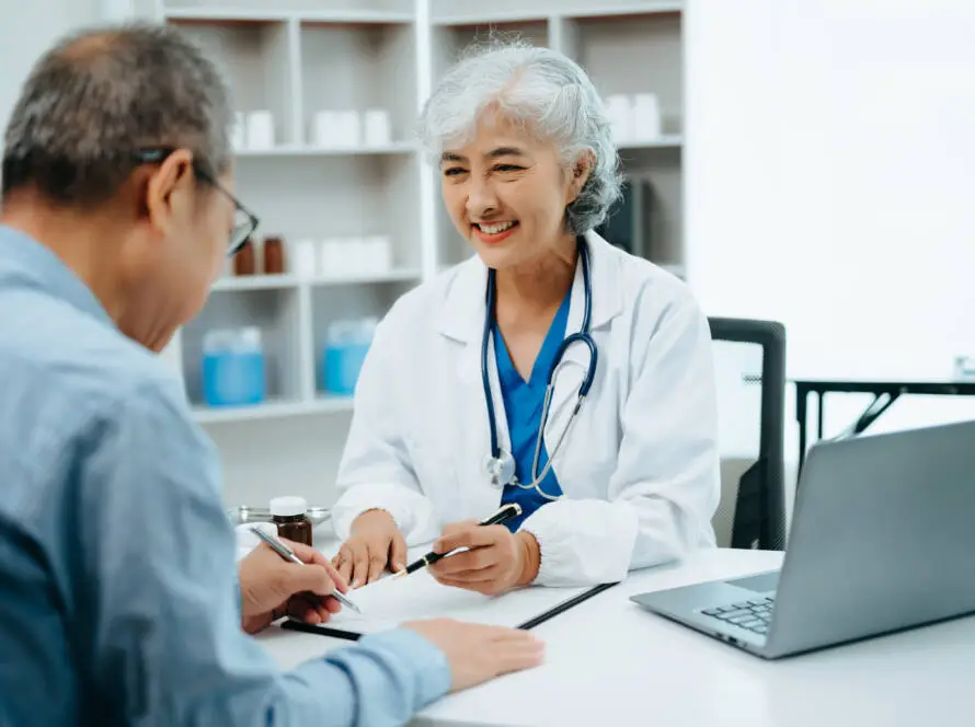 An older female doctor with grey hair and a stethoscope smiles while talking to a male patient across the desk. She gestures to a clipboard, highlighting personalised care—a focus for any top Healthcare Marketing Agency.