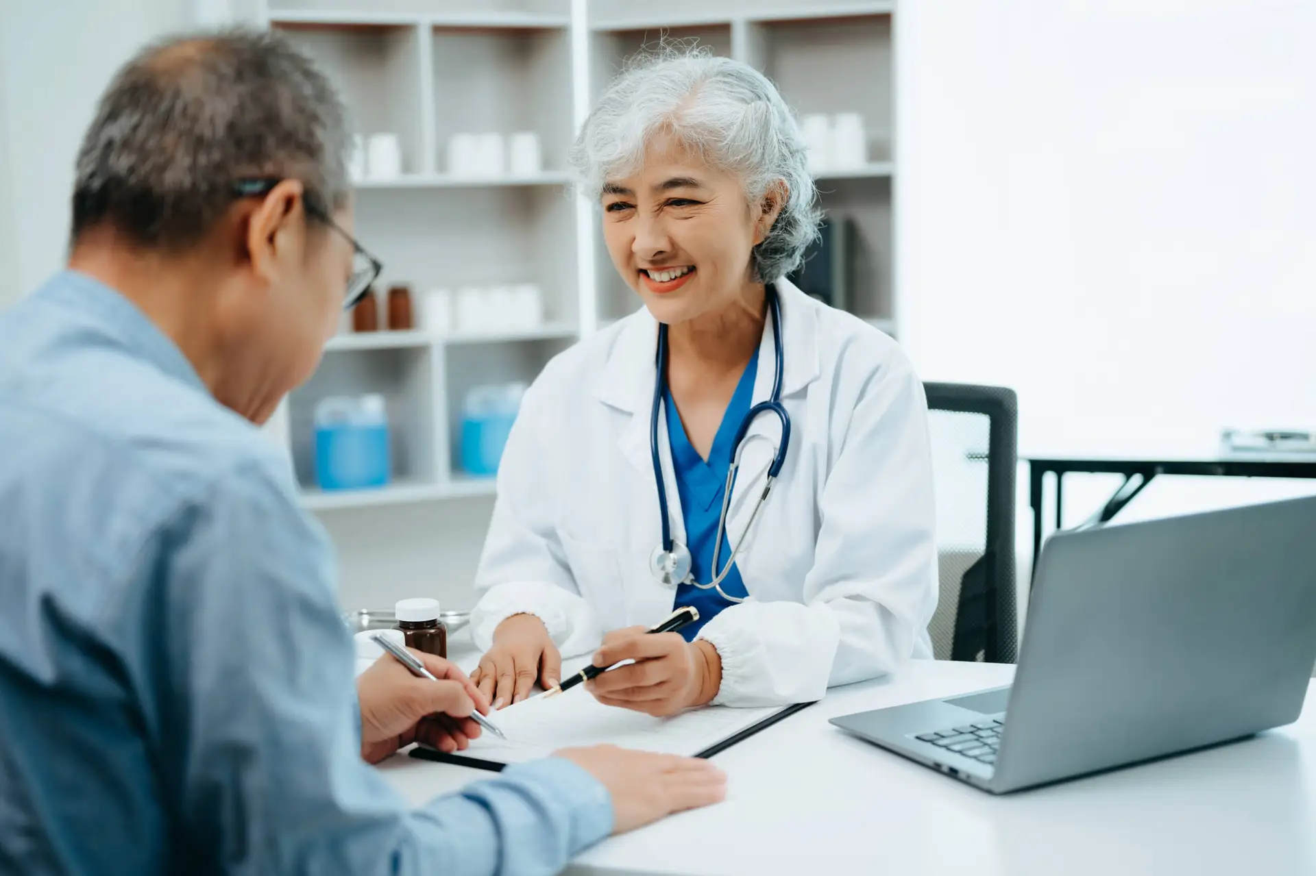 An older female doctor with grey hair and a stethoscope smiles while talking to a male patient across the desk. She gestures to a clipboard, highlighting personalised care—a focus for any top Healthcare Marketing Agency.