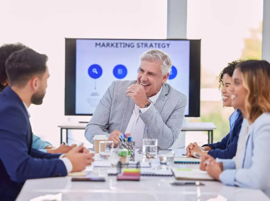 Five business professionals sit around a conference table, smiling and engaged in discussion at a Healthcare Marketing Agency. A man in a light grey suit laughs at the centre while a Marketing Strategy presentation is displayed on the screen.