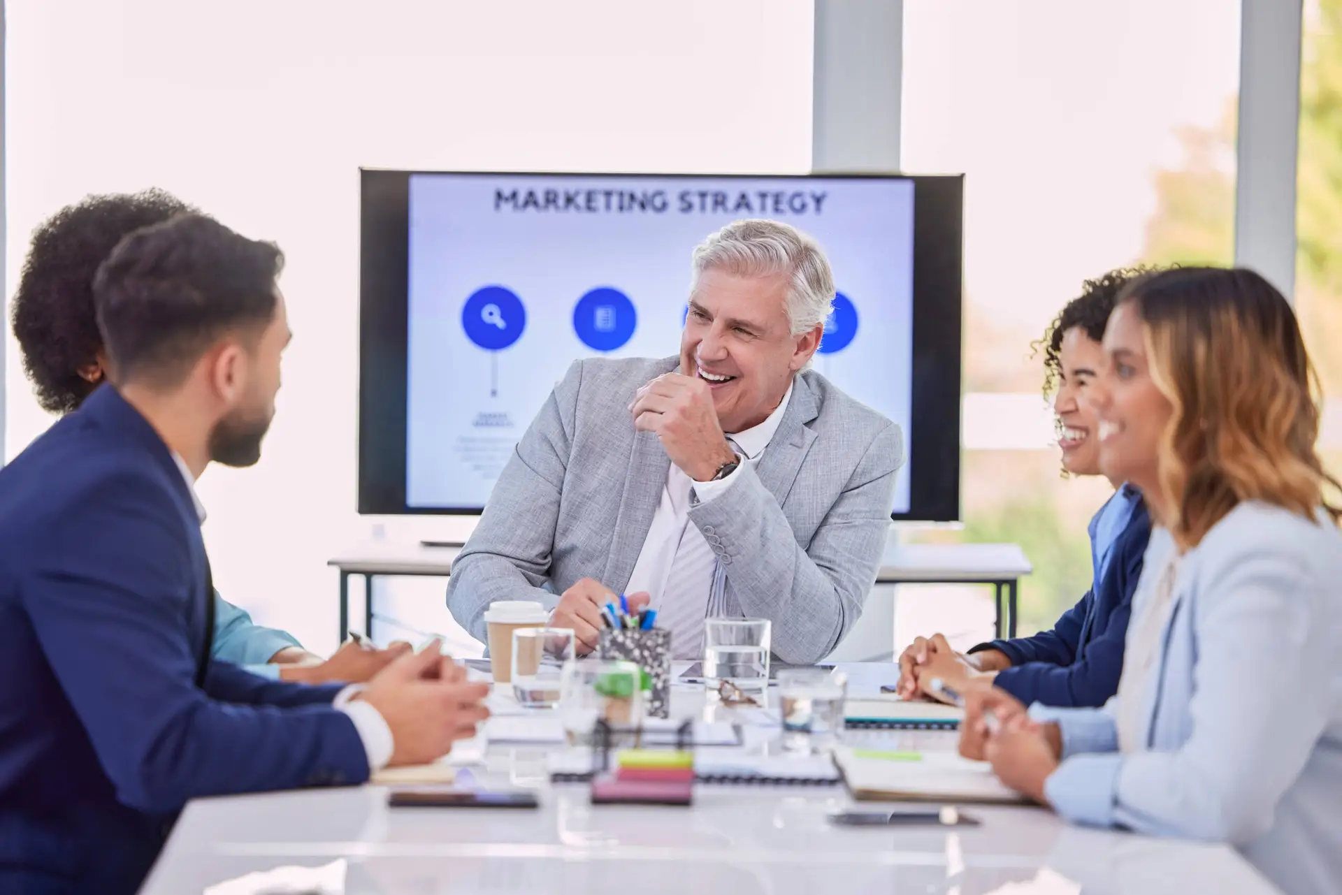 Five business professionals sit around a conference table, smiling and engaged in discussion at a Healthcare Marketing Agency. A man in a light grey suit laughs at the centre while a Marketing Strategy presentation is displayed on the screen.