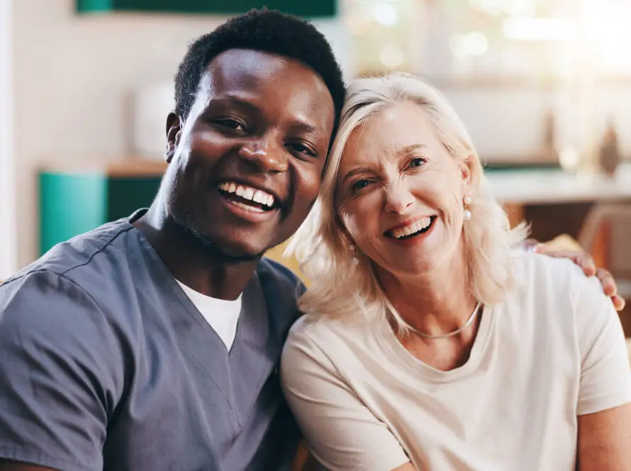 A smiling male healthcare worker in scrubs sits arm-in-arm with a smiling older woman in a bright, homely living room, conveying warmth and support—ideal imagery for a Healthcare Marketing Agency focused on compassionate care.