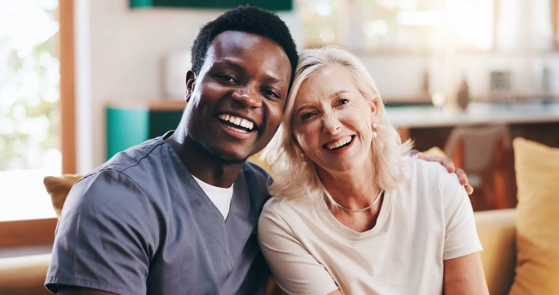 A smiling male healthcare worker in scrubs sits arm-in-arm with a smiling older woman in a bright, homely living room, conveying warmth and support—ideal imagery for a Healthcare Marketing Agency focused on compassionate care.