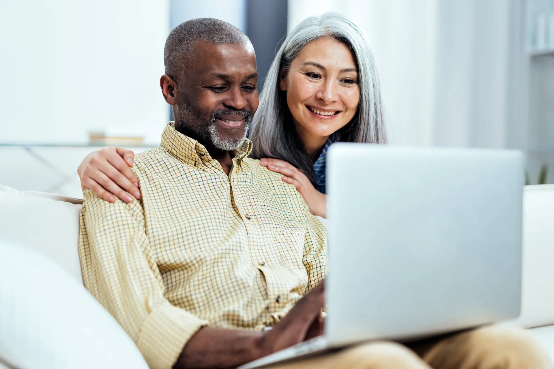 A smiling older couple sit closely together on a sofa, looking at a laptop. Their happy, relaxed expressions suggest they're exploring options with a Healthcare Marketing Agency in their bright, cosy room.