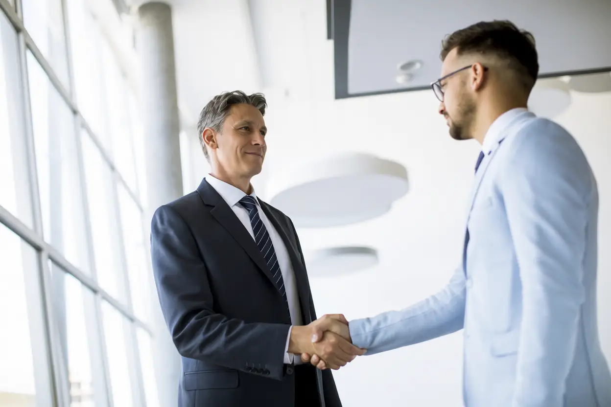 Two men in business suits shake hands in a bright, modern office with large windows. The scene suggests a professional agreement, possibly sealing a partnership with a leading Healthcare Marketing Agency.