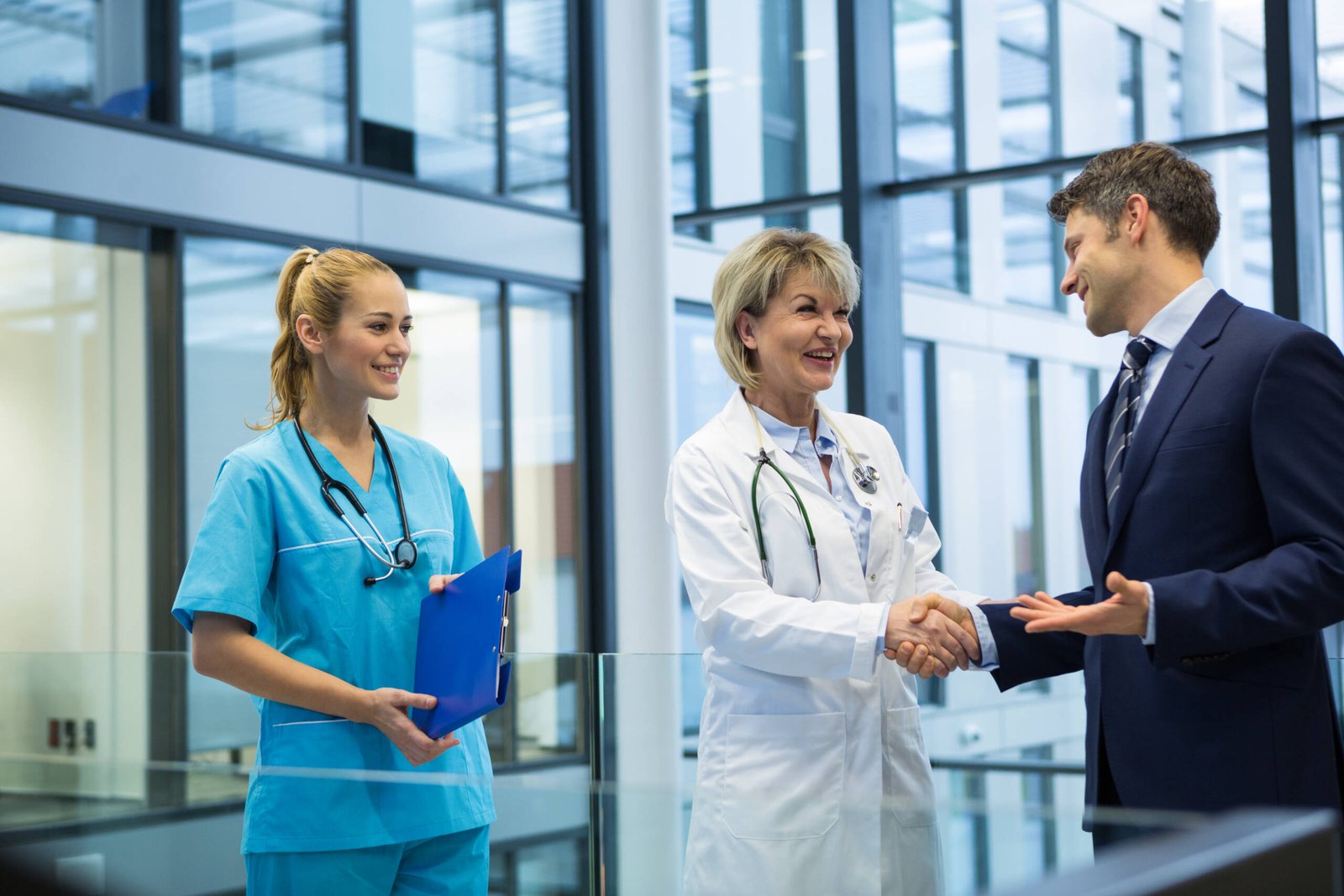 A female doctor in a white coat shakes hands with a man in a business suit, both smiling. Next to them, a nurse in blue scrubs holds a clipboard. They stand in a bright hospital lobby, representing a Weight Loss Clinics Marketing Agency partnership.
