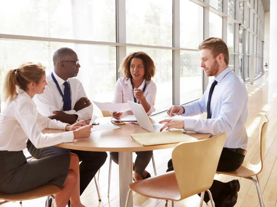 Four professionals sit around a round table in a bright, modern office. Three have laptops and documents on the table, discussing strategies for a Healthcare Marketing Agency. One woman speaks as natural light fills the room and trees are visible outside.