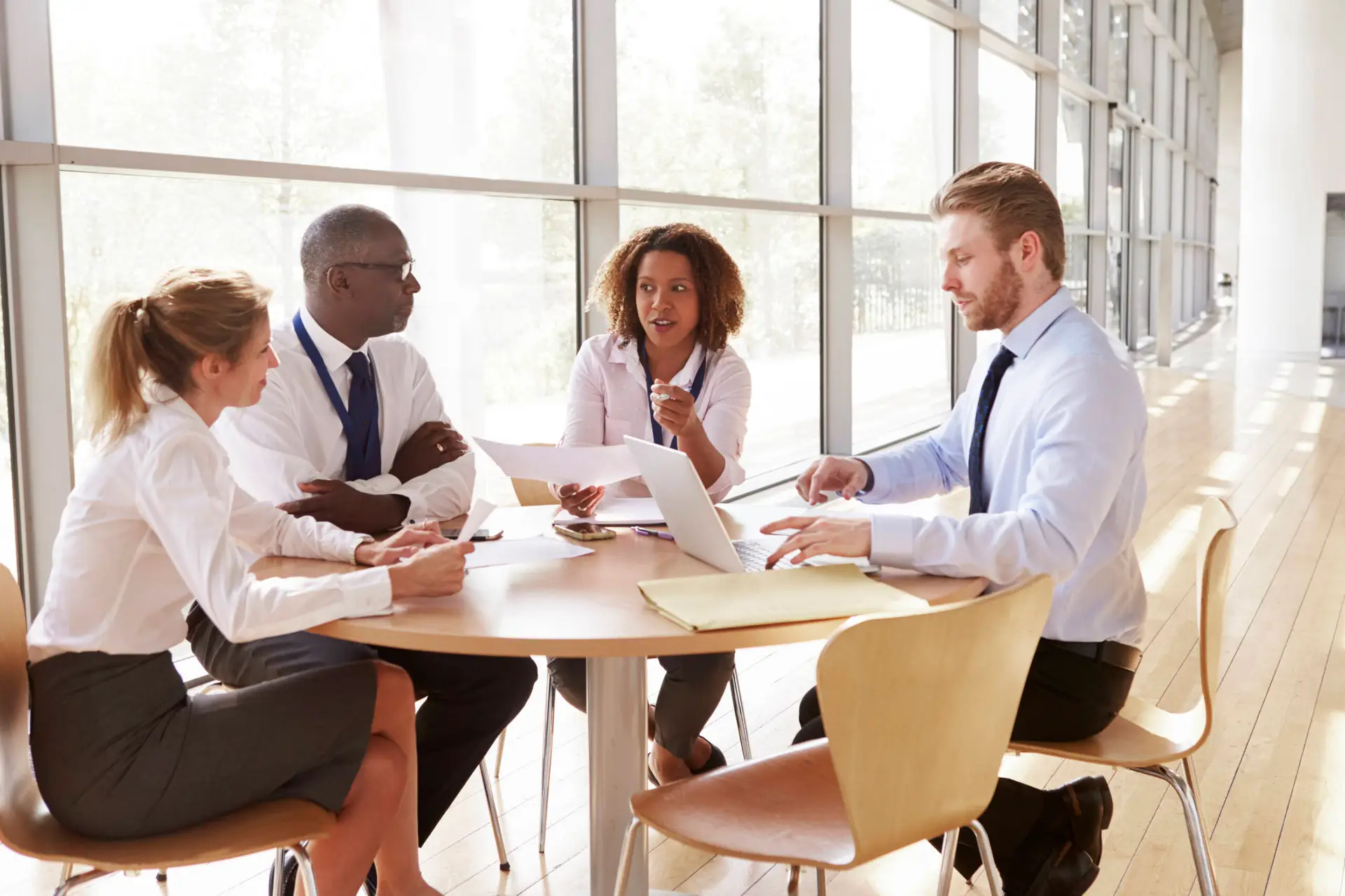 Four professionals sit around a round table in a bright, modern office. Three have laptops and documents on the table, discussing strategies for a Healthcare Marketing Agency. One woman speaks as natural light fills the room and trees are visible outside.
