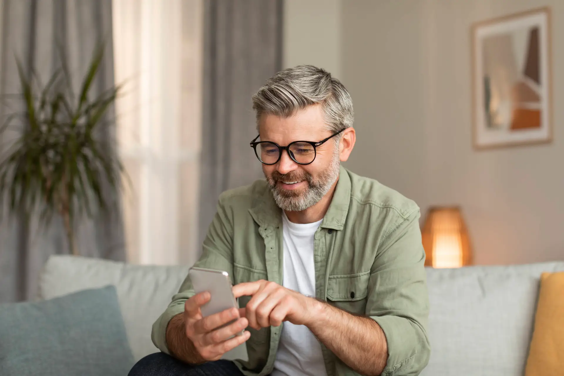 A smiling middle-aged man with grey hair and beard, wearing glasses, a green shirt, and a white T-shirt, sits on a light sofa in a cosy living room. He is browsing on his mobile phone, possibly exploring a healthcare marketing agency online.
