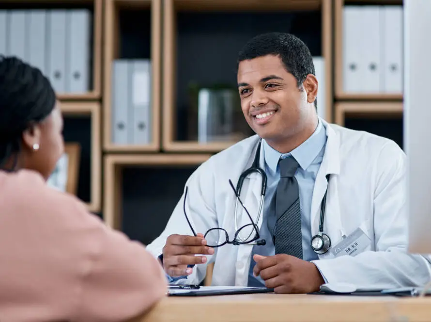 A male doctor in a white coat and stethoscope smiles and holds spectacles while discussing treatment options with a female patient in a modern surgery—an ideal setting highlighted by any top healthcare marketing agency.