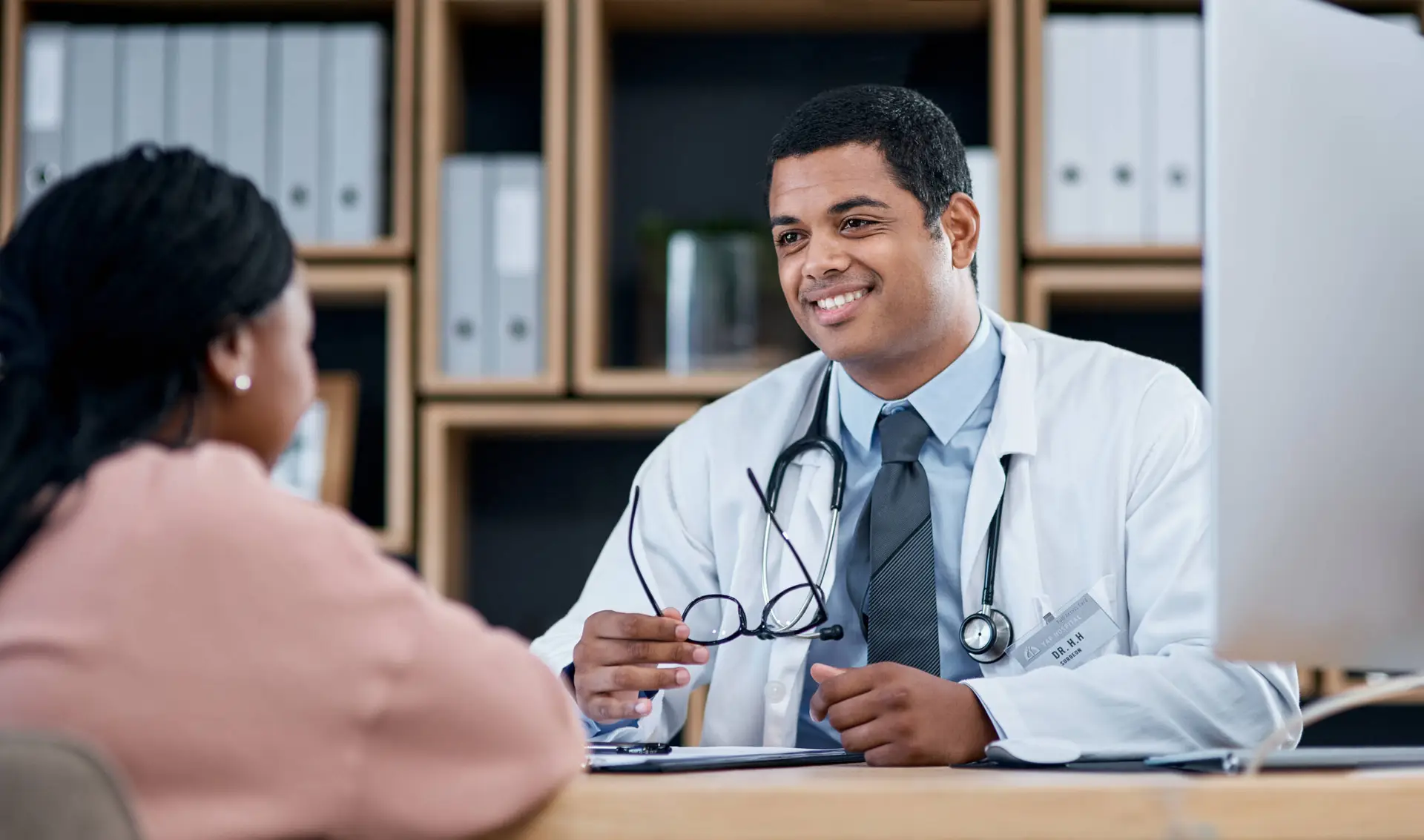 A male doctor in a white coat and stethoscope smiles and holds spectacles while discussing treatment options with a female patient in a modern surgery—an ideal setting highlighted by any top healthcare marketing agency.