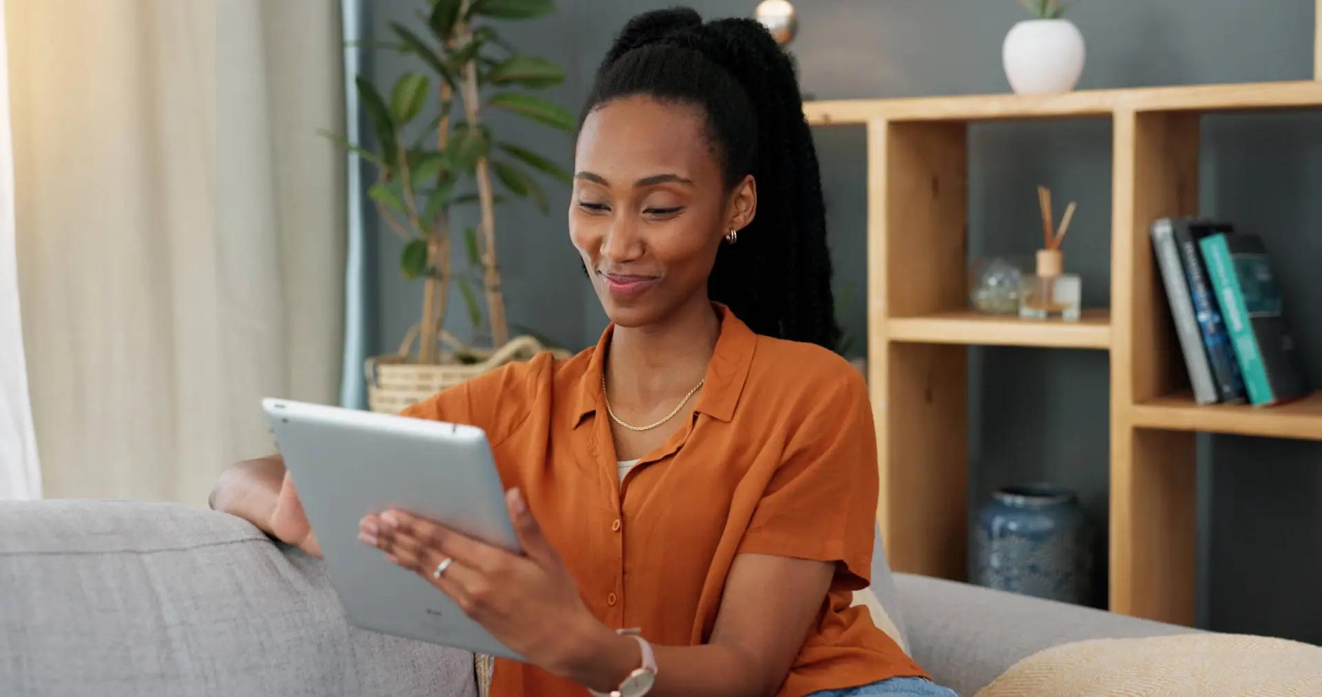 A woman with long dark hair in a ponytail, wearing an orange shirt and gold jewellery, sits on a grey sofa smiling while looking at a tablet—perhaps reviewing insights from a Healthcare Marketing Agency. A wooden shelf with books and plants is behind her.