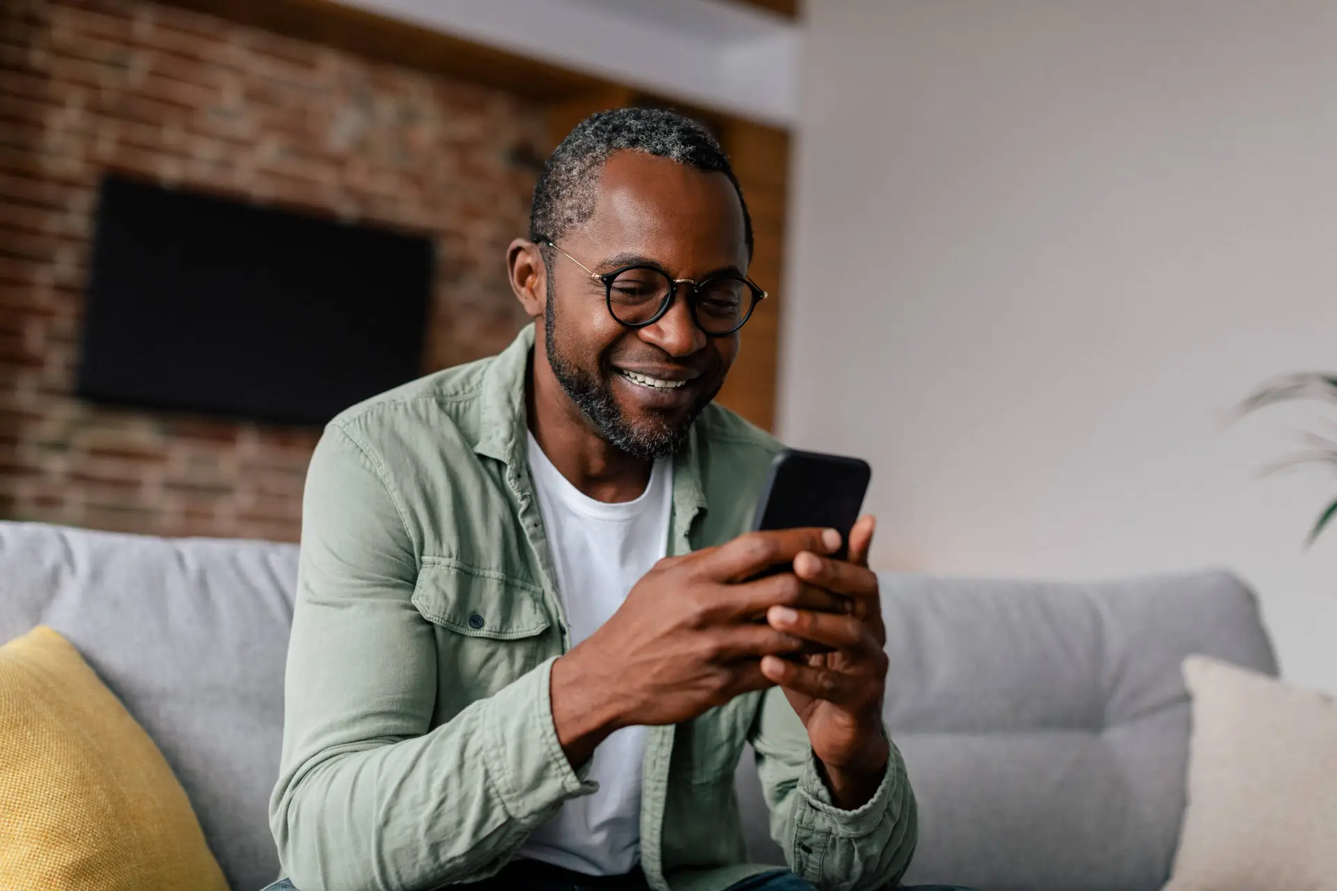 A smiling man with short hair, glasses, and a beard sits on a grey sofa with yellow and grey cushions. He wears a green shirt over a white T-shirt and browses his mobile for the latest tips from a Healthcare Marketing Agency. A brick wall and television are in the background.