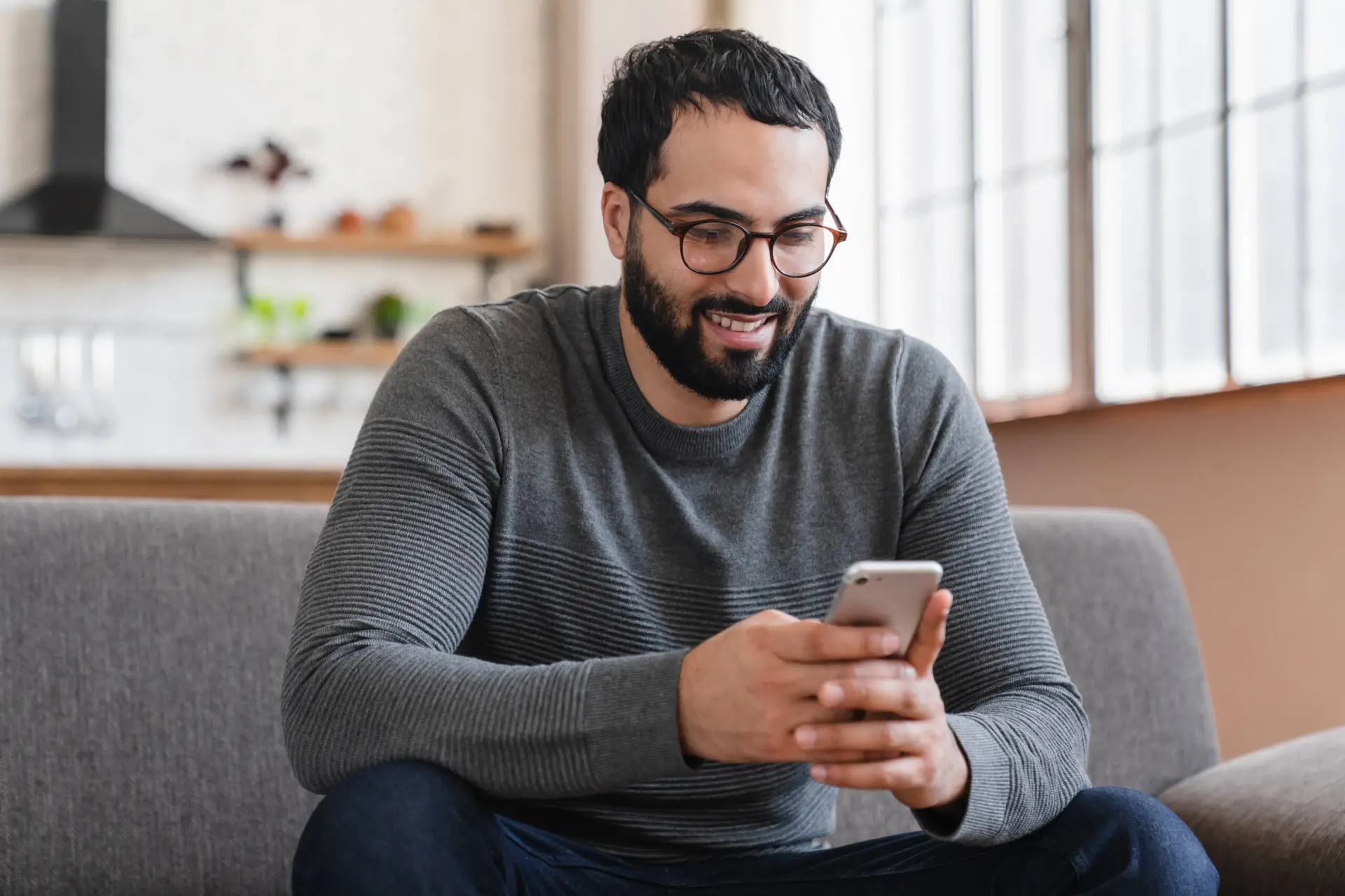 A man with dark hair, a beard, and glasses sits on a grey sofa, smiling while looking at his mobile—perhaps exploring ideas for a Healthcare Marketing Agency. Large windows and a bright, modern interior appear in the background.