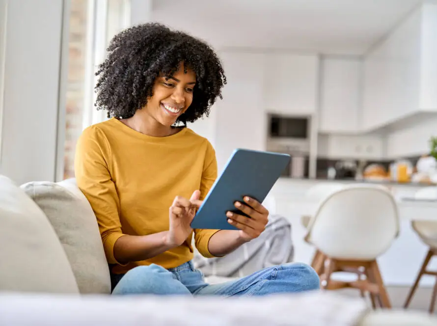 A woman with curly hair, wearing a mustard yellow top and jeans, sits cross-legged on a sofa, smiling whilst browsing a Healthcare Marketing Agency’s website on her tablet in a bright, modern kitchen and living room space.