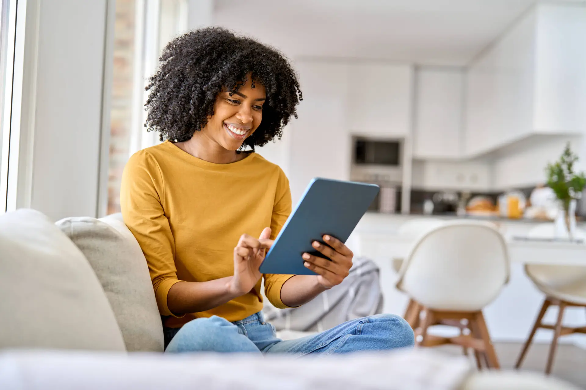 A woman with curly hair, wearing a mustard yellow top and jeans, sits cross-legged on a sofa, smiling whilst browsing a Healthcare Marketing Agency’s website on her tablet in a bright, modern kitchen and living room space.