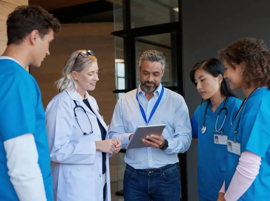 A doctor in a white coat and three healthcare workers in blue scrubs stand with a man holding a tablet. In this modern medical facility, they appear attentive and engaged, perhaps consulting with a Hair Transplant Clinics Marketing Agency.