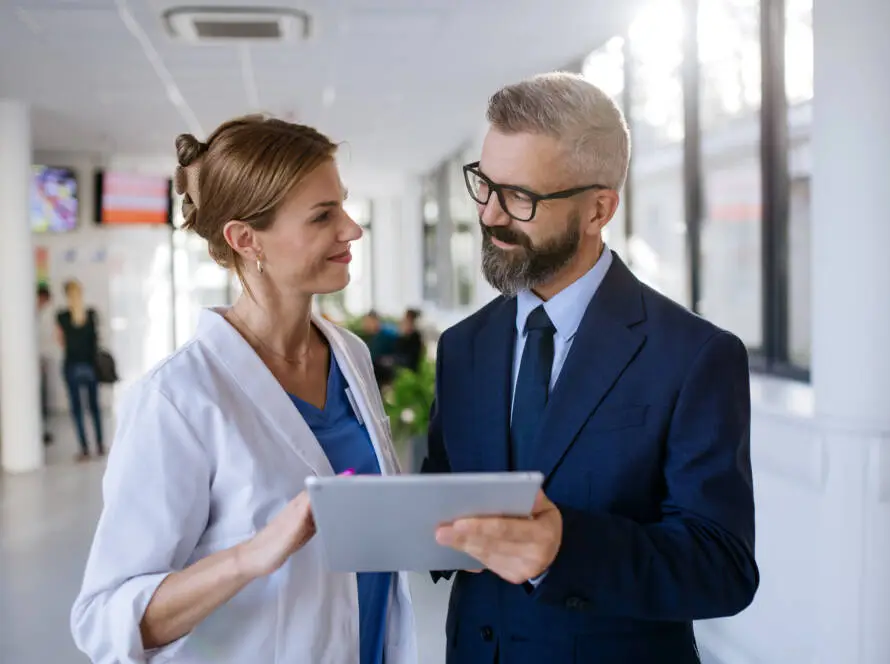 A female doctor in a white coat holds a tablet and smiles whilst talking to a man in a navy suit and glasses. They stand in a modern corridor, reflecting the professional environment of a leading healthcare marketing agency.