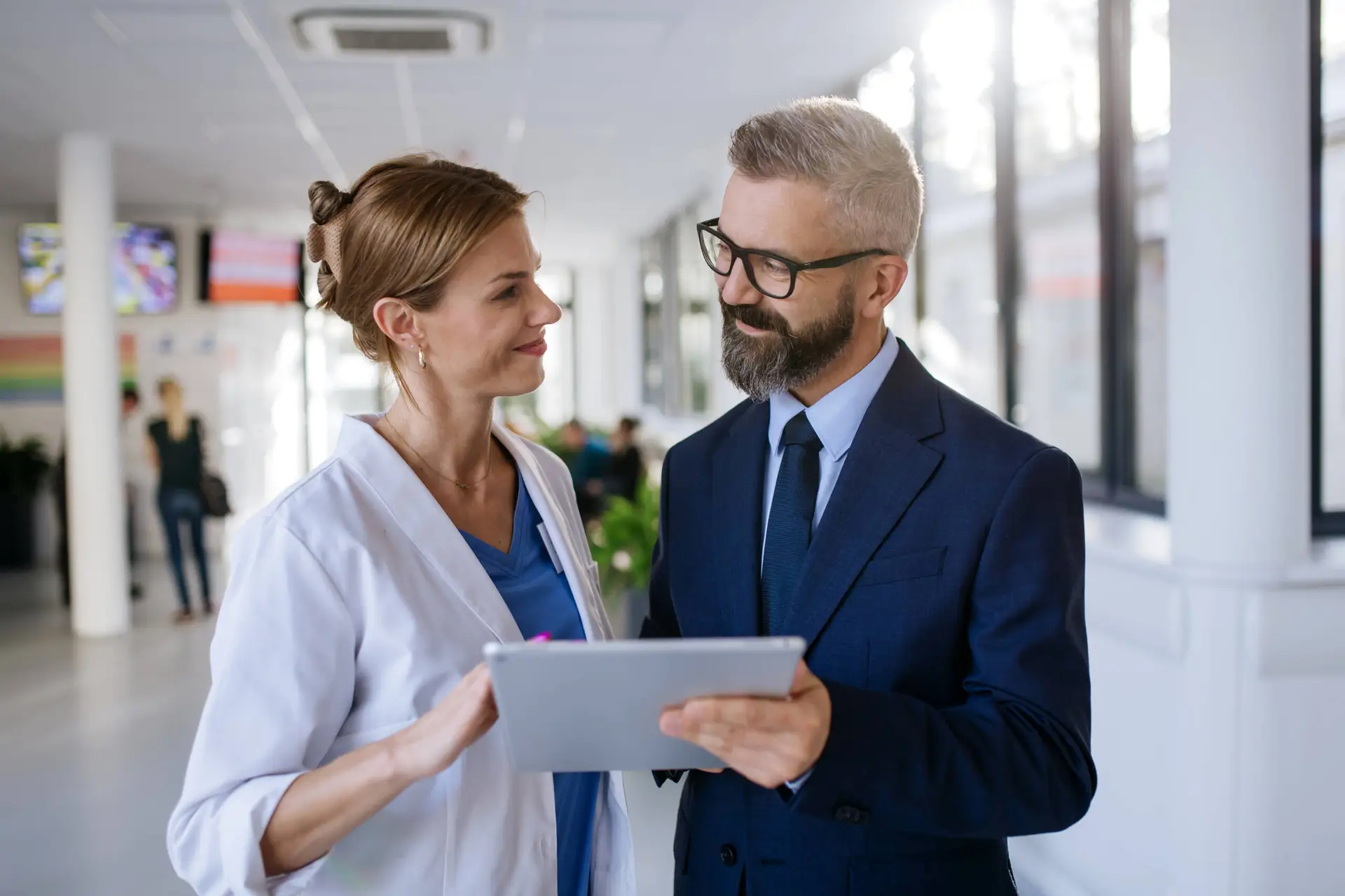 A female doctor in a white coat holds a tablet and smiles whilst talking to a man in a navy suit and glasses. They stand in a modern corridor, reflecting the professional environment of a leading healthcare marketing agency.