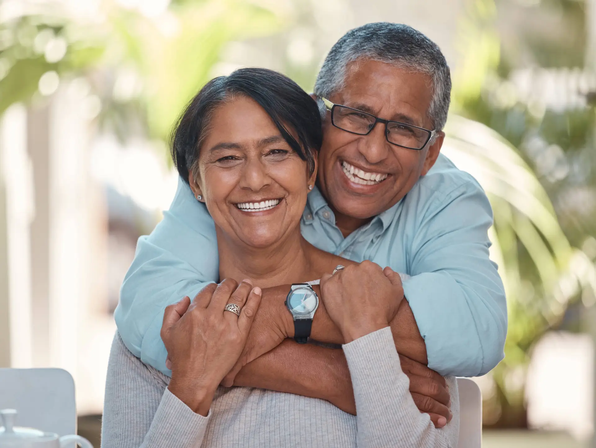 An older couple smiles warmly at the camera. The man hugs her shoulders as they relax in a sunlit setting with green plants blurred behind—capturing a moment of happiness that a Healthcare Marketing Agency might use to promote wellbeing and connection.