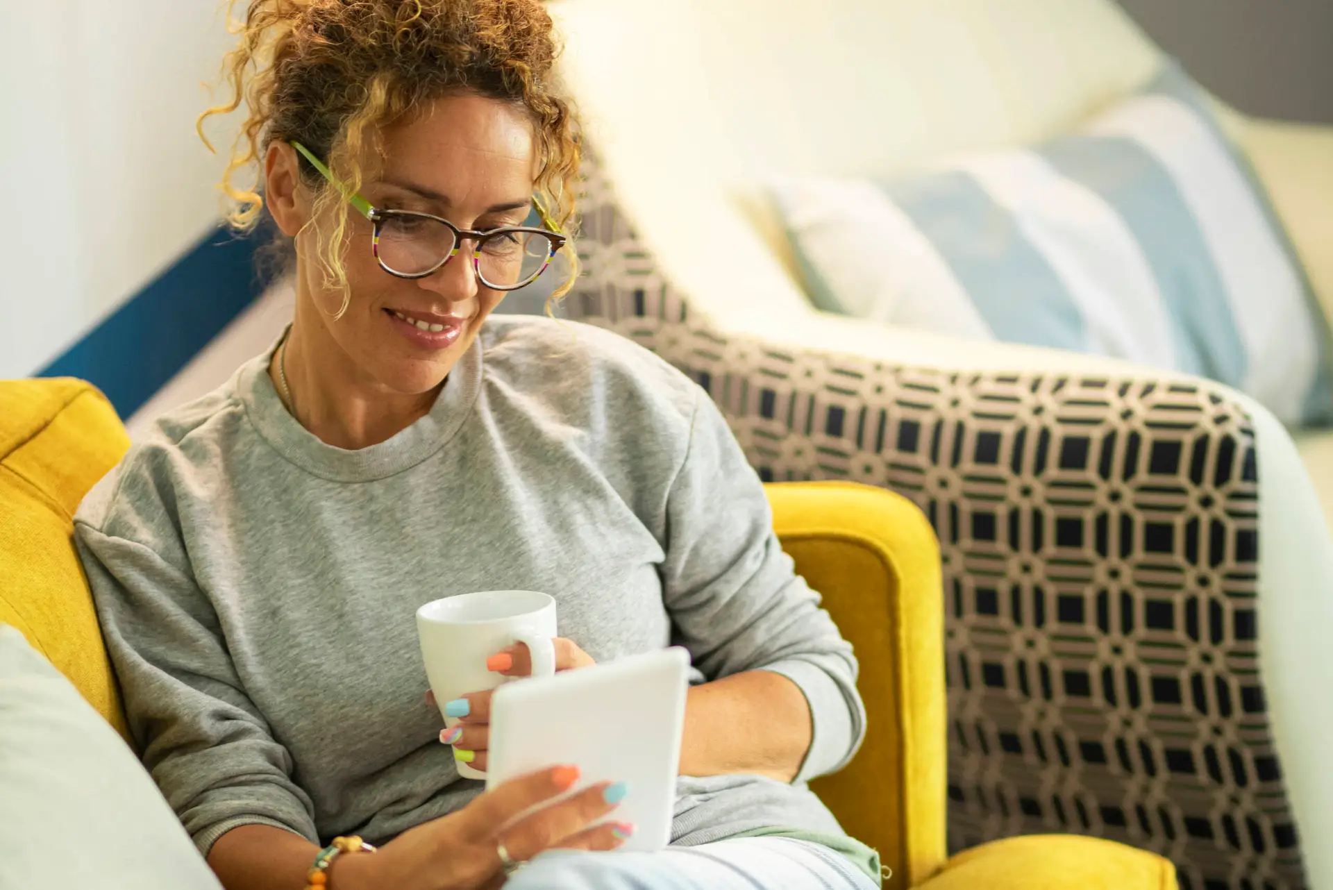 A woman with curly hair and glasses sits on a yellow sofa, smiling while holding a white mug and a tablet—perhaps catching up on the latest from her favourite healthcare marketing agency. She wears a grey sweatshirt, with patterned cushions in the background.