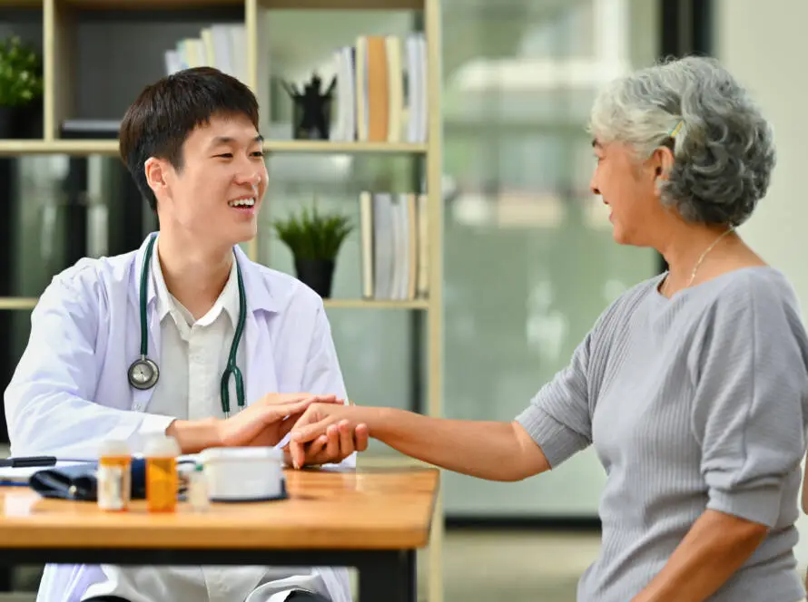 A smiling male doctor in a white coat, representing a healthcare marketing agency, checks the pulse of an older woman across a wooden desk with medicine bottles and devices. Shelves lined with books and plants add warmth to the scene.