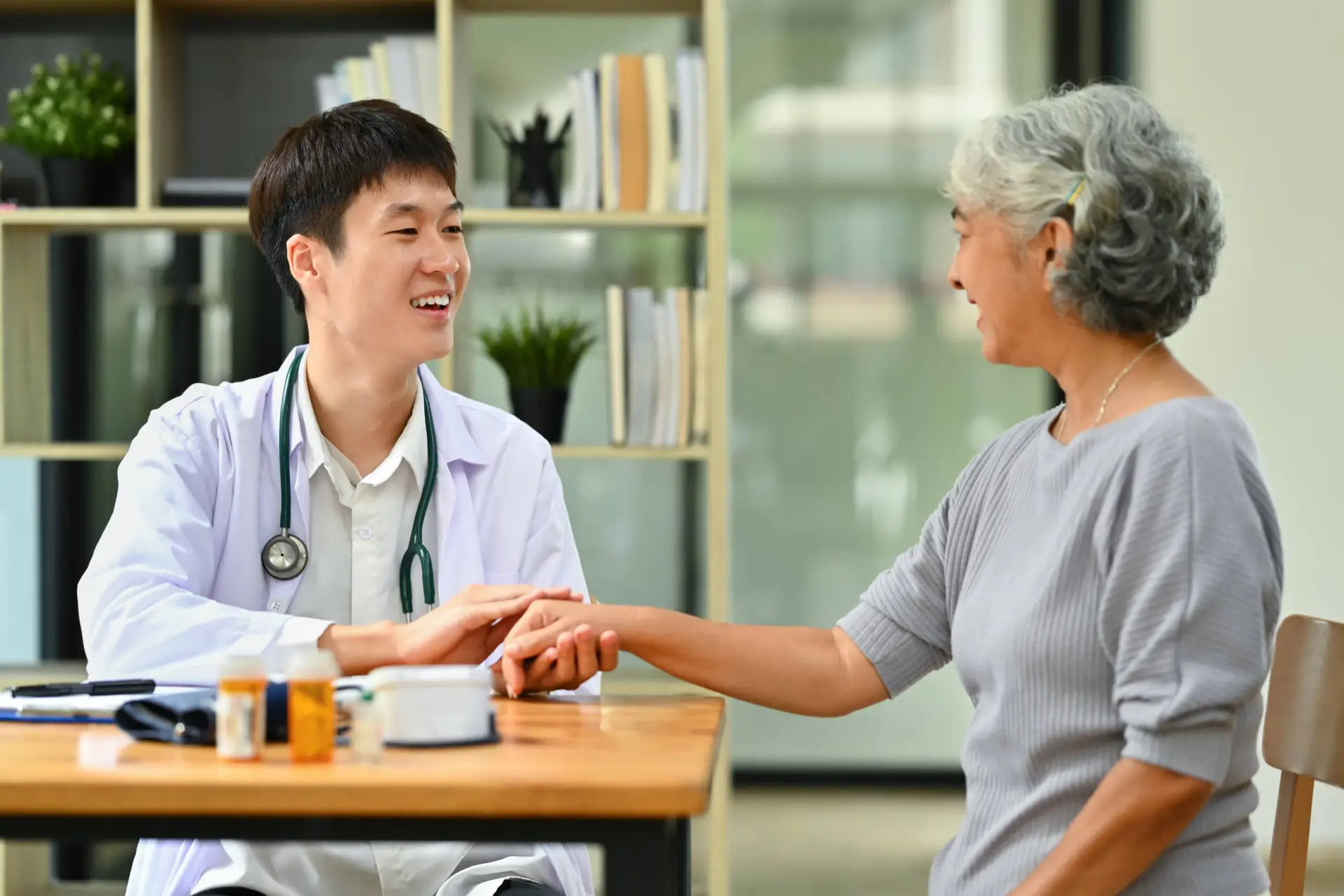 A smiling male doctor in a white coat, representing a healthcare marketing agency, checks the pulse of an older woman across a wooden desk with medicine bottles and devices. Shelves lined with books and plants add warmth to the scene.