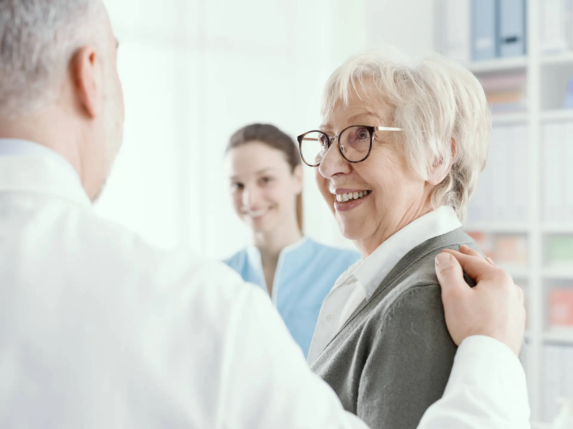 An older woman with short grey hair and glasses smiles at a male doctor who reassures her. A young female nurse in blue scrubs stands in the background, also smiling. The scene reflects compassionate care—a value championed by any leading healthcare marketing agency.
