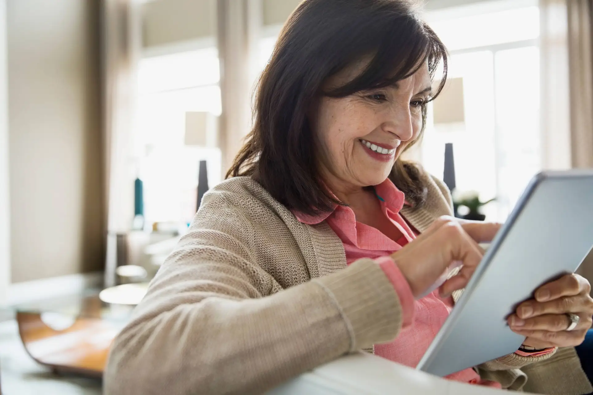 A smiling woman with dark hair, wearing a light brown cardigan and pink shirt, sits indoors using a tablet. She appears relaxed and focused, perhaps reviewing insights from a leading healthcare marketing agency as natural light streams in behind her.
