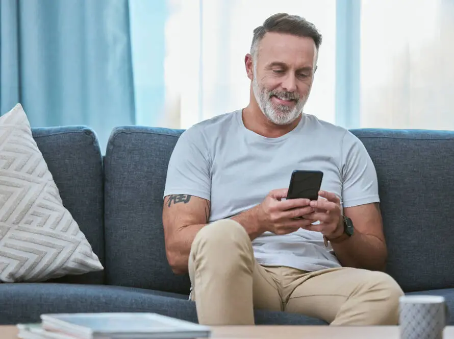A middle-aged man with short grey hair and a beard sits on a blue sofa, smiling at his mobile—perhaps reading tips from a Healthcare Marketing Agency. He wears a light grey T-shirt and beige trousers, showing off a tattoo on his right arm.