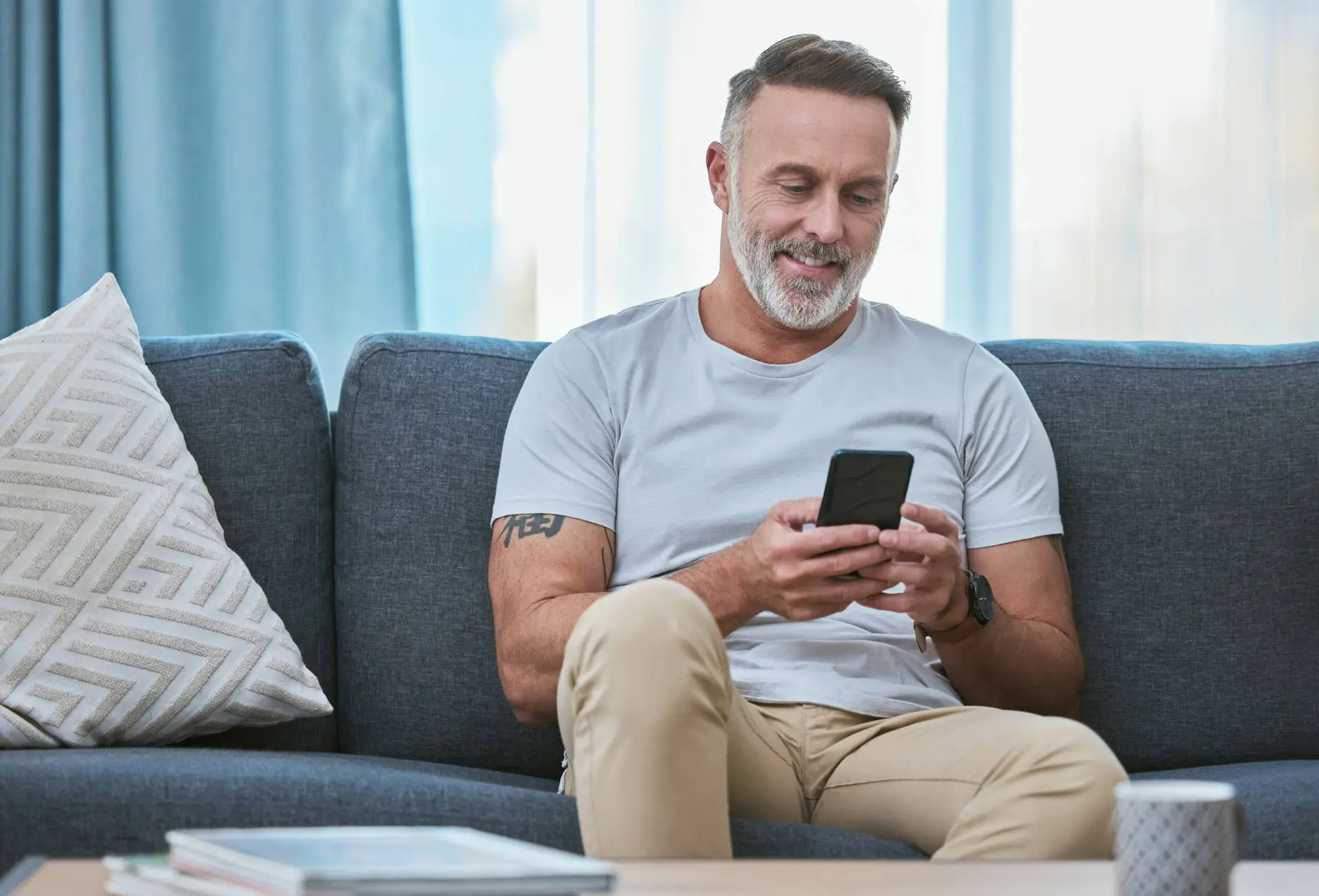 A middle-aged man with short grey hair and a beard sits on a blue sofa, smiling at his mobile—perhaps reading tips from a Healthcare Marketing Agency. He wears a light grey T-shirt and beige trousers, showing off a tattoo on his right arm.