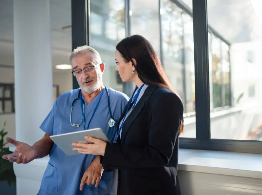 A male doctor in blue scrubs and a stethoscope discusses healthcare strategies with a woman from a Healthcare Marketing Agency, who holds a tablet. They stand by large windows in a bright corridor, sunlight illuminating their faces.