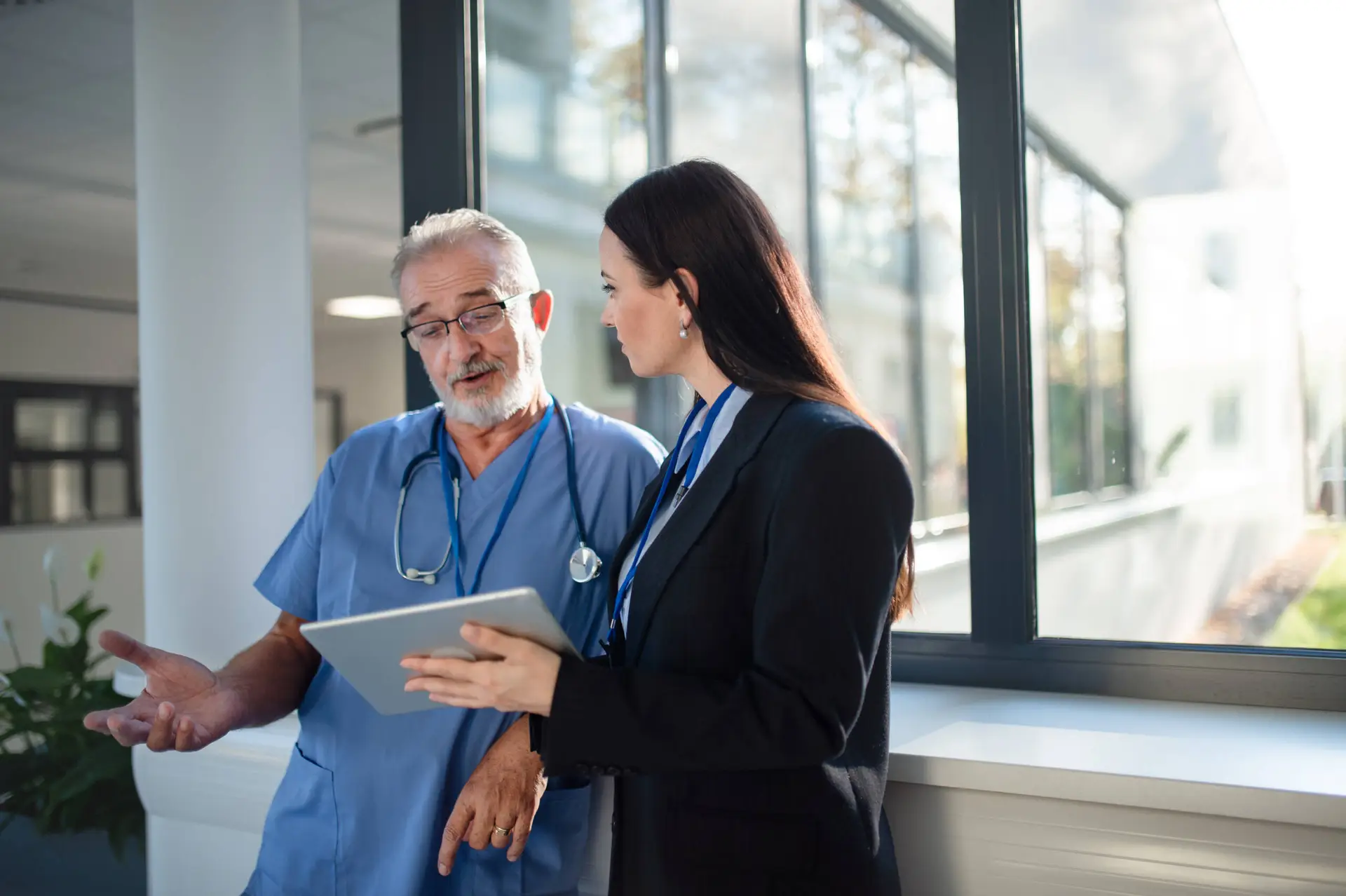 A male doctor in blue scrubs and a stethoscope discusses healthcare strategies with a woman from a Healthcare Marketing Agency, who holds a tablet. They stand by large windows in a bright corridor, sunlight illuminating their faces.