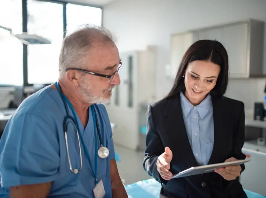A male doctor in blue scrubs with a stethoscope sits beside a woman in a black suit. Smiling, she shows him something on her tablet—an example of how a Healthcare Marketing Agency can support modern medical practices in a bright, contemporary office.