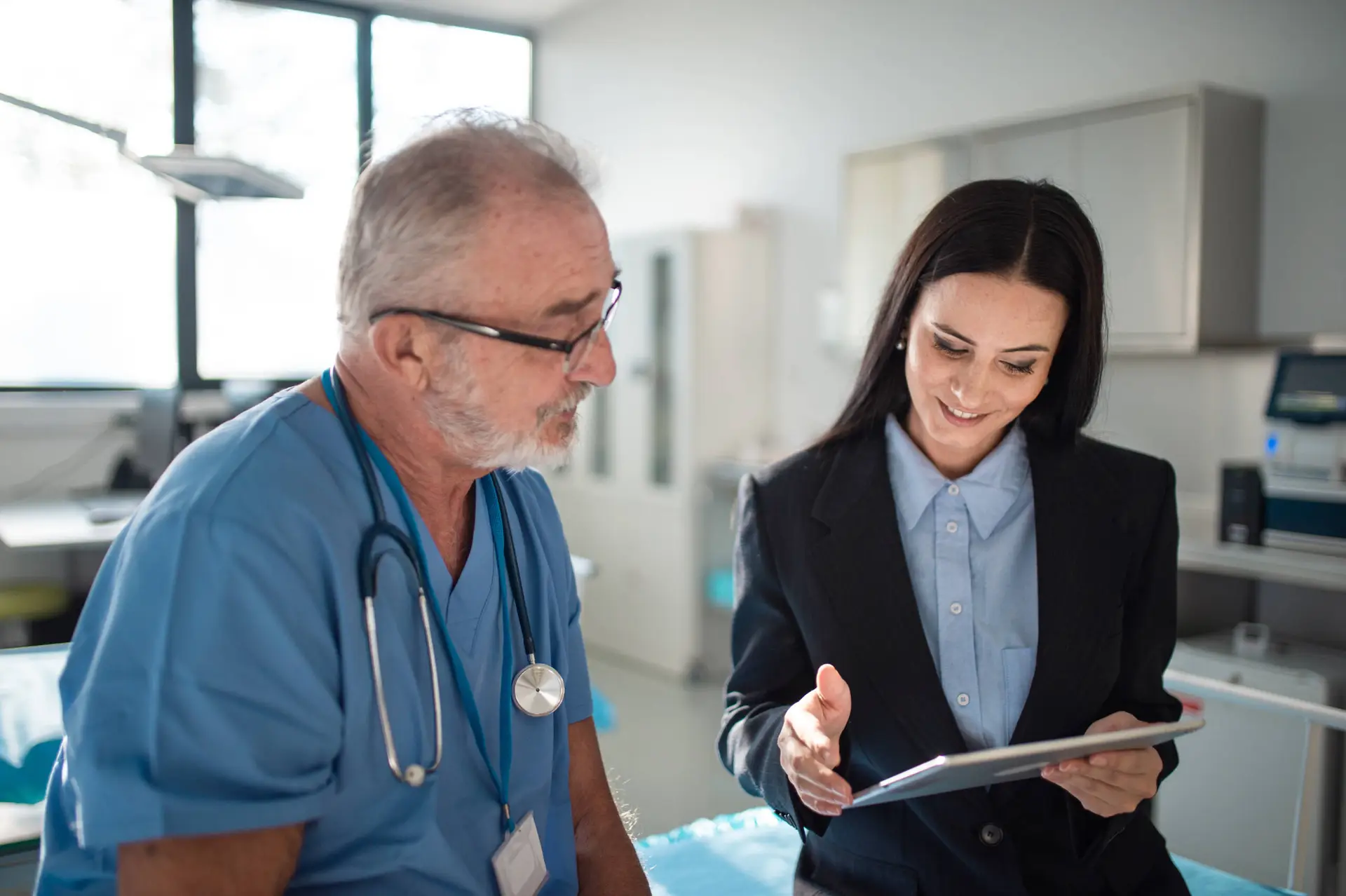 A male doctor in blue scrubs with a stethoscope sits beside a woman in a black suit. Smiling, she shows him something on her tablet—an example of how a Healthcare Marketing Agency can support modern medical practices in a bright, contemporary office.