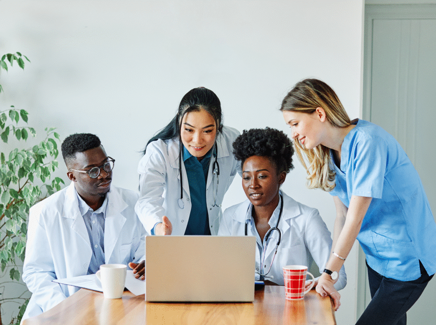 Four healthcare professionals, including two women in white coats, a man in a white coat, and a woman in blue scrubs, gather round a laptop at a wooden table. They appear to be discussing something on the screen. Cups and papers are on the table.