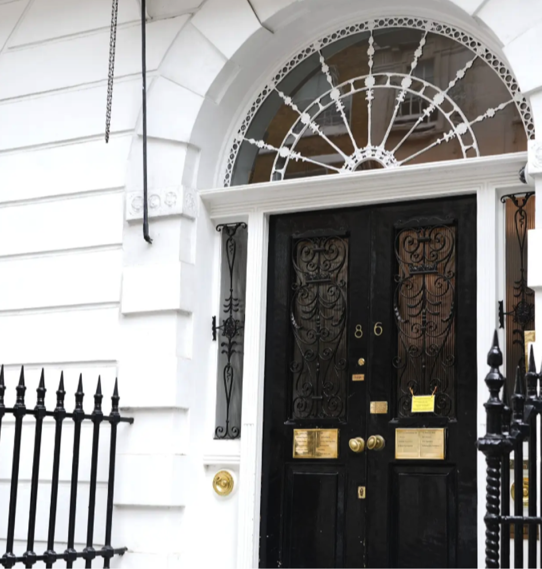 A black double door with ornate ironwork and brass letterboxes is set in a white building—a striking entrance worthy of architectural case studies. The semicircular fanlight features decorative glass, and number 86 is displayed above black railings and a white wall.