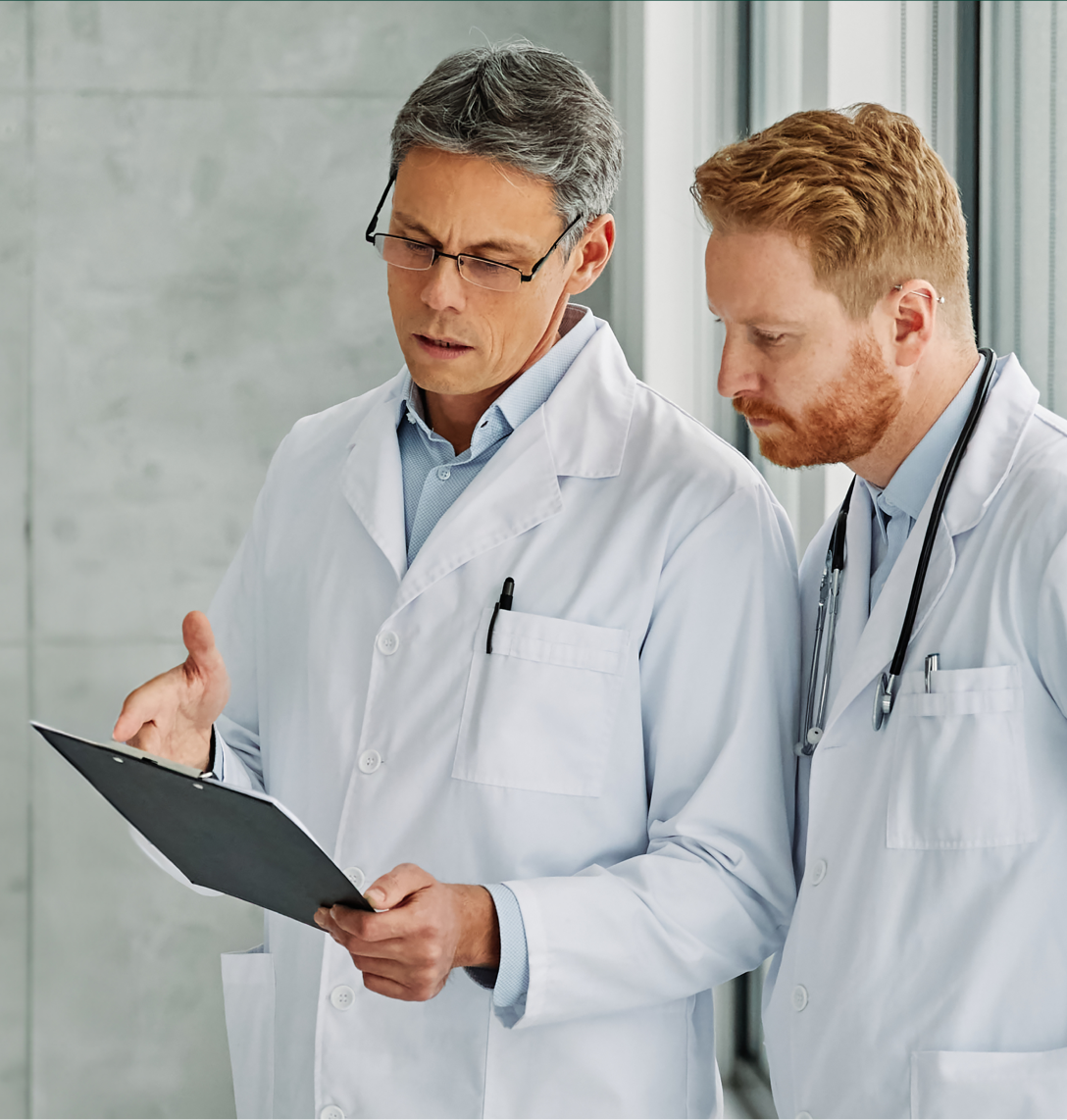 Two male doctors in white lab coats stand indoors by a window, closely reviewing and discussing case studies on a clipboard. One has grey hair and spectacles; the other has red hair and a stethoscope. Both appear focused and serious.