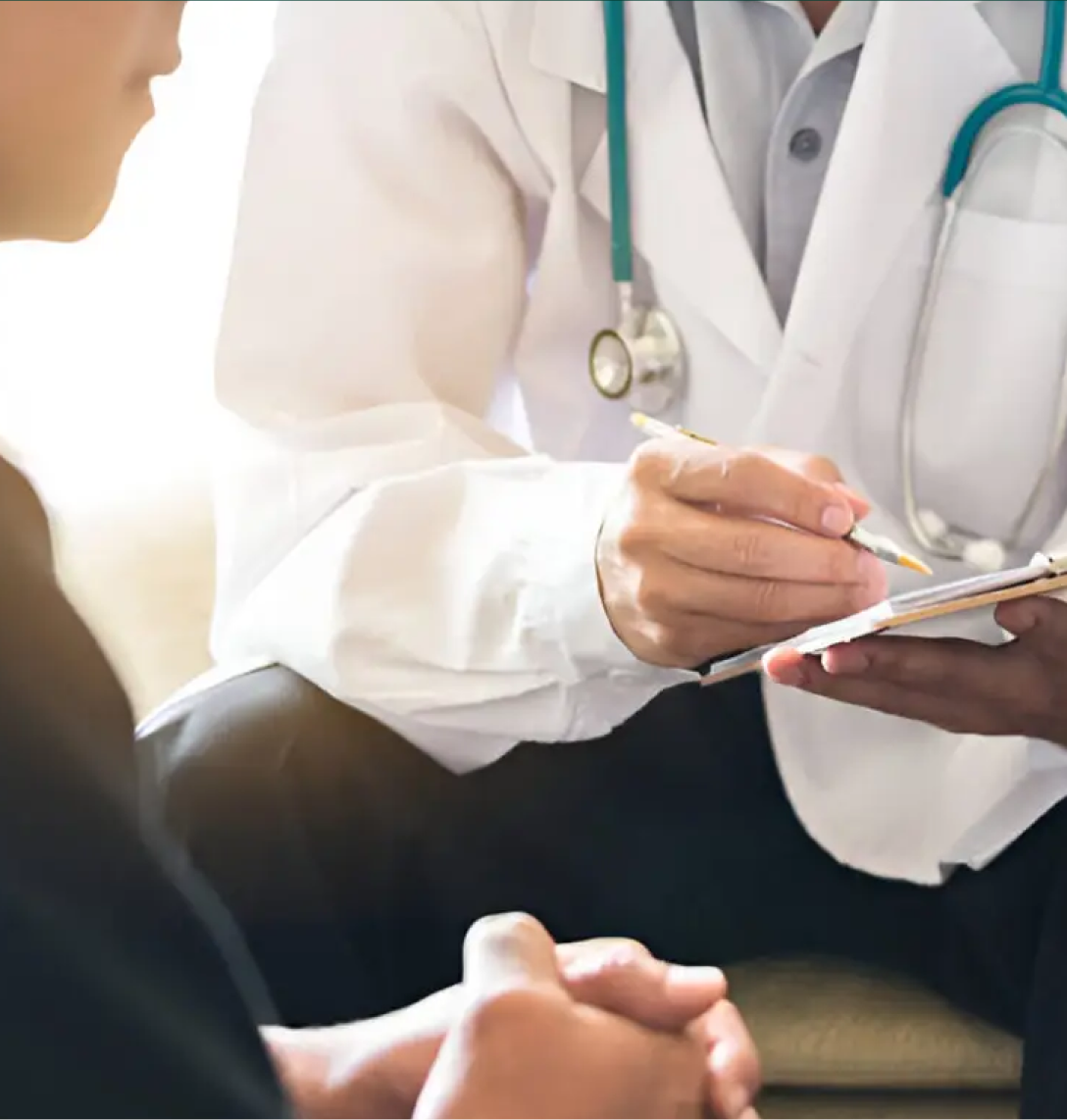 A doctor in a white coat with a stethoscope holds a clipboard and pen, discussing case studies with a seated patient. The patient’s clasped hands and visible torso suggest an in-depth consultation in a well-lit room.