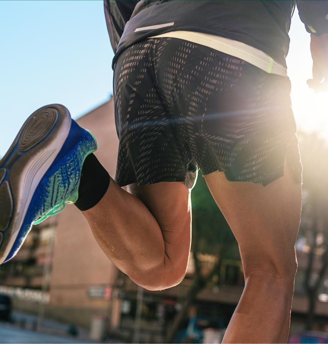 Close-up view from behind of a person running outdoors in sunlight, wearing patterned shorts, a jacket, and blue running trainers—like many featured in fitness case studies—with cityscape and trees visible in the background.