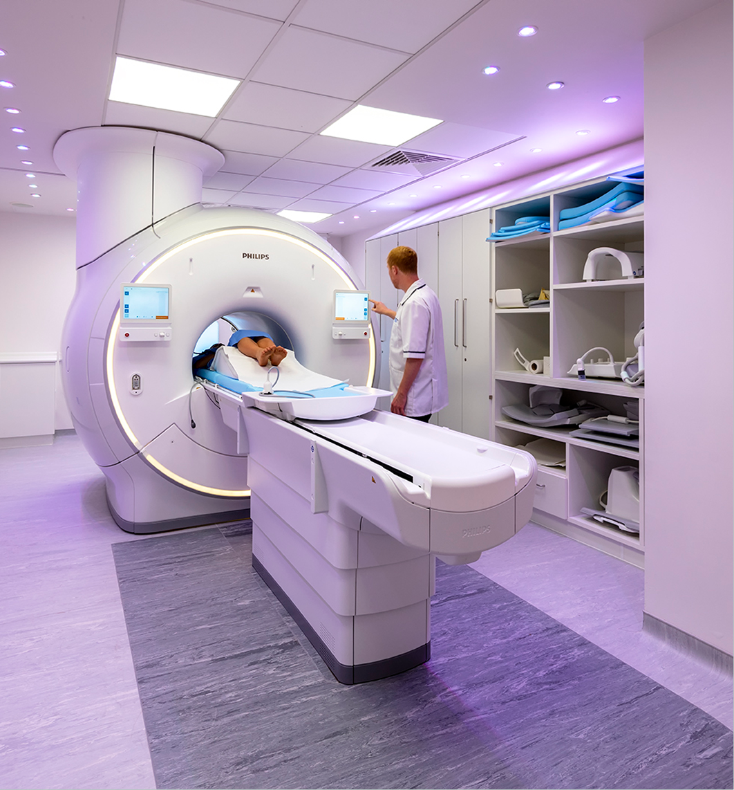 A patient lies on a sliding table entering a large, white MRI machine. A healthcare worker operates a touchscreen panel nearby. Shelves with medical accessories and case studies line the wall. The room is brightly lit with purple accent lights.