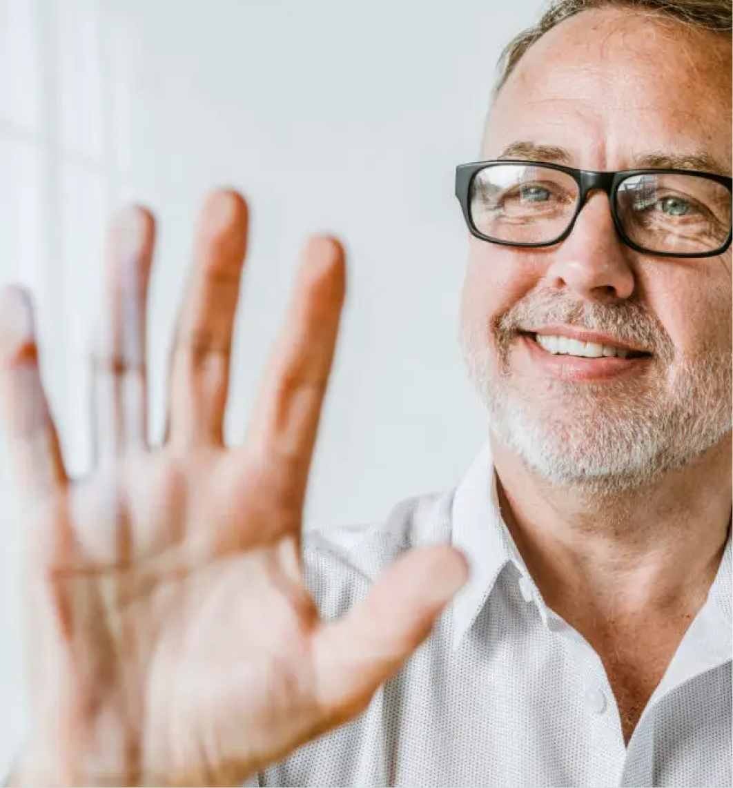 A middle-aged man with glasses and a grey beard smiles warmly, holding his right hand up towards a glass surface—like he's ready to discuss compelling case studies. He wears a light-coloured shirt, with natural light brightening the clean, white background.